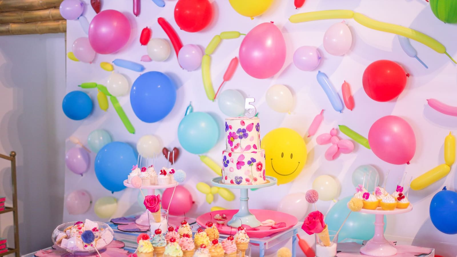 A colourful kids party dessert table with a floral cake and a wall of balloons
