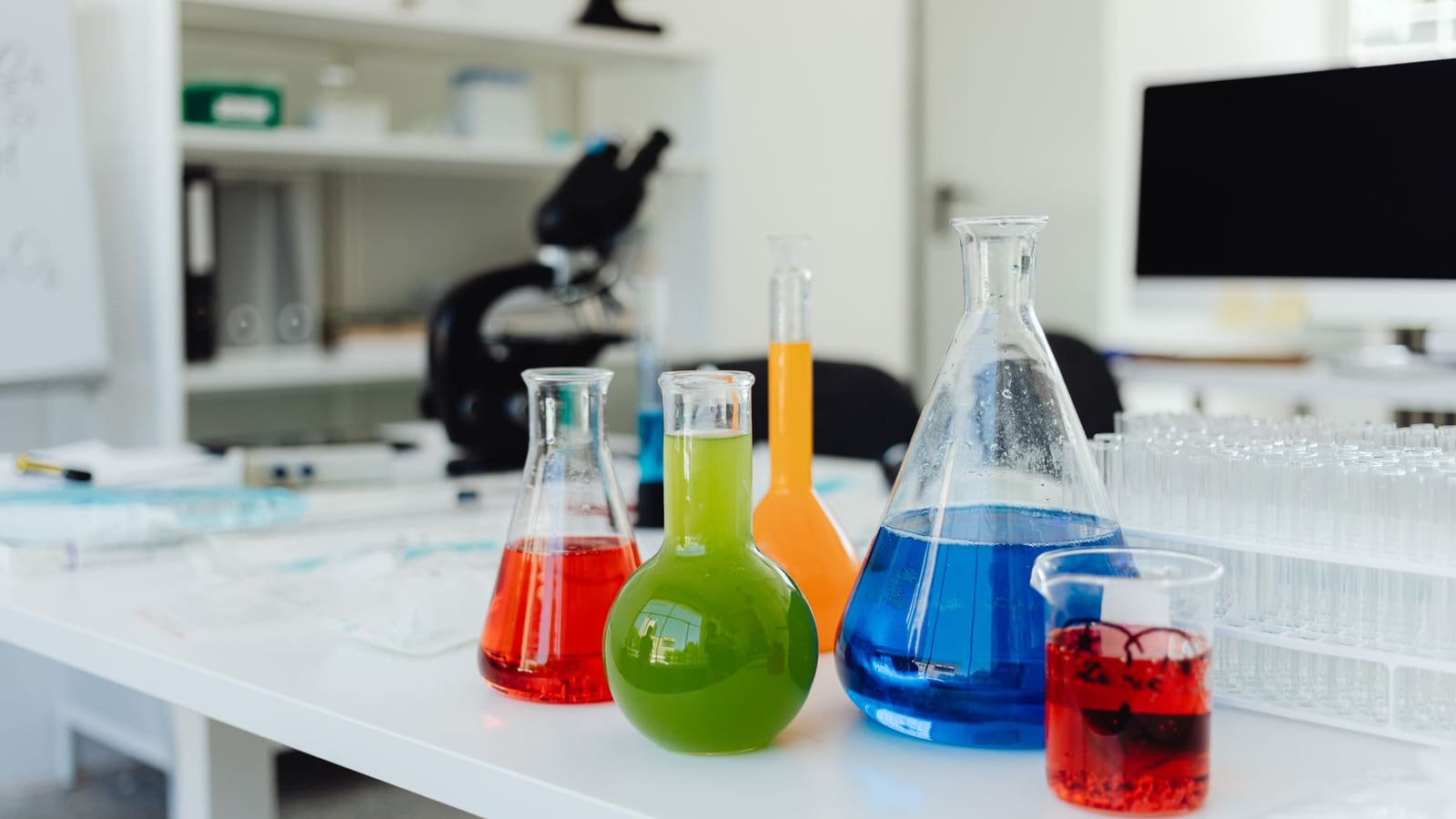 Conical flasks of bright red, green, orange and blue liquid on a bench with a microscope behind